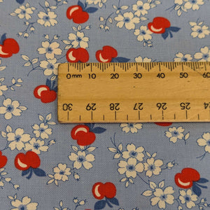 Close up of fabric with red apples and white flowers on a blue background in a vintage style, for sale at Minerva's Bower with a ruler included for scale